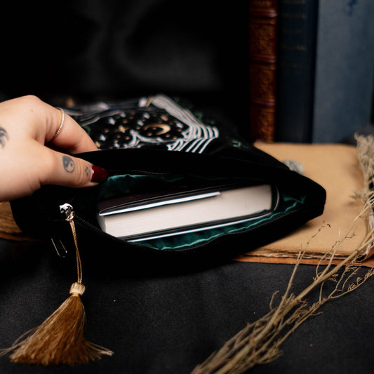 Hand opening a black velvet book pouch with a tassel on a dark surface with books and dried plants.