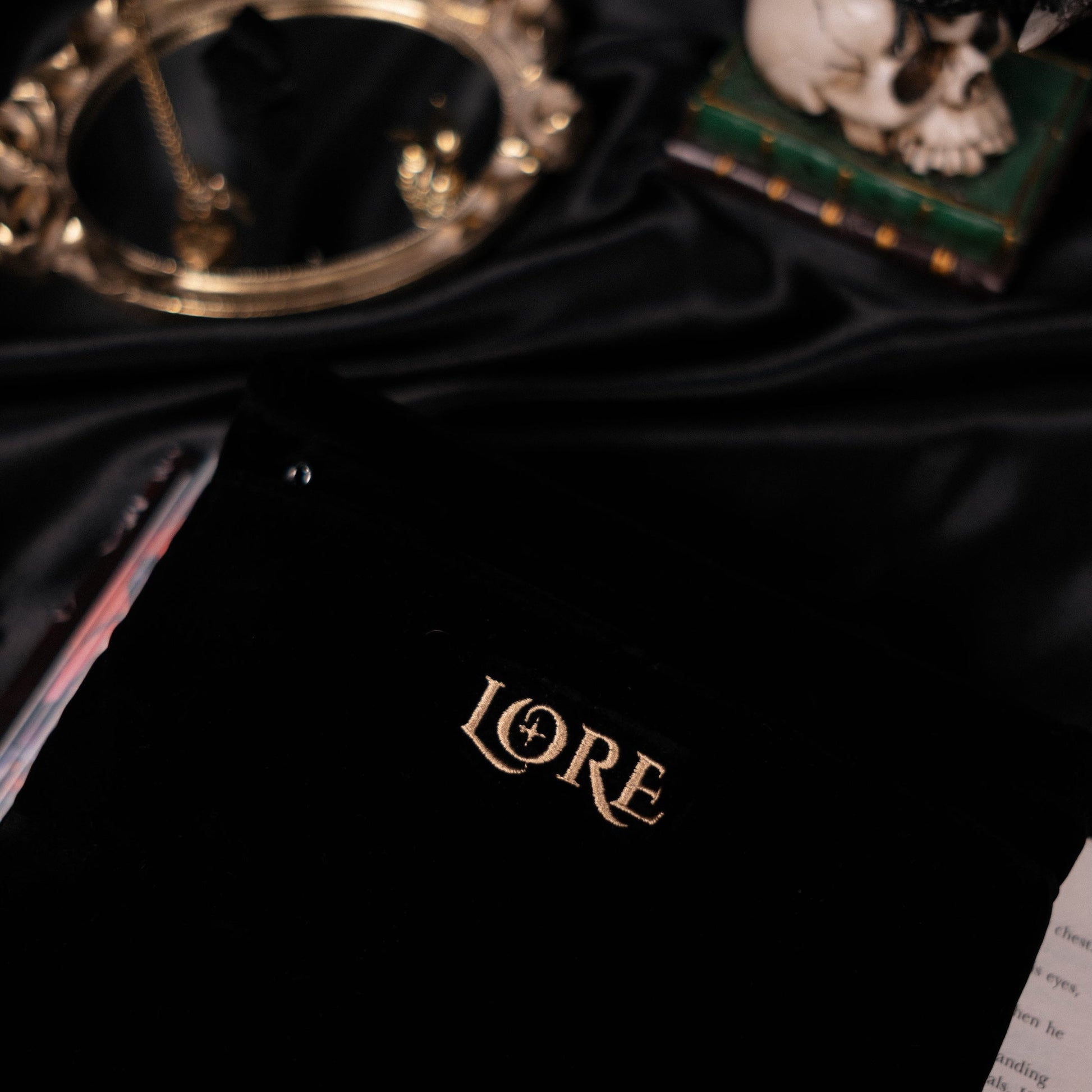 Black book sleeve with 'LORE' logo in gold lettering, accompanied by a gold mirror and skull on a dark fabric background.