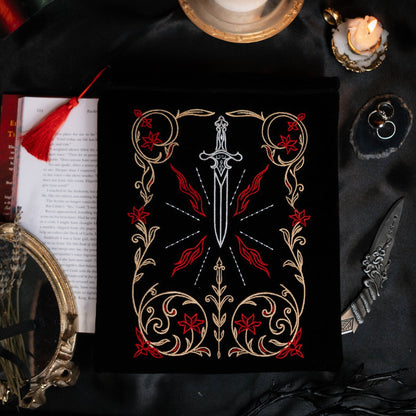 Decorative book sleeve with a sword design on a dark fabric background with candles and books.
