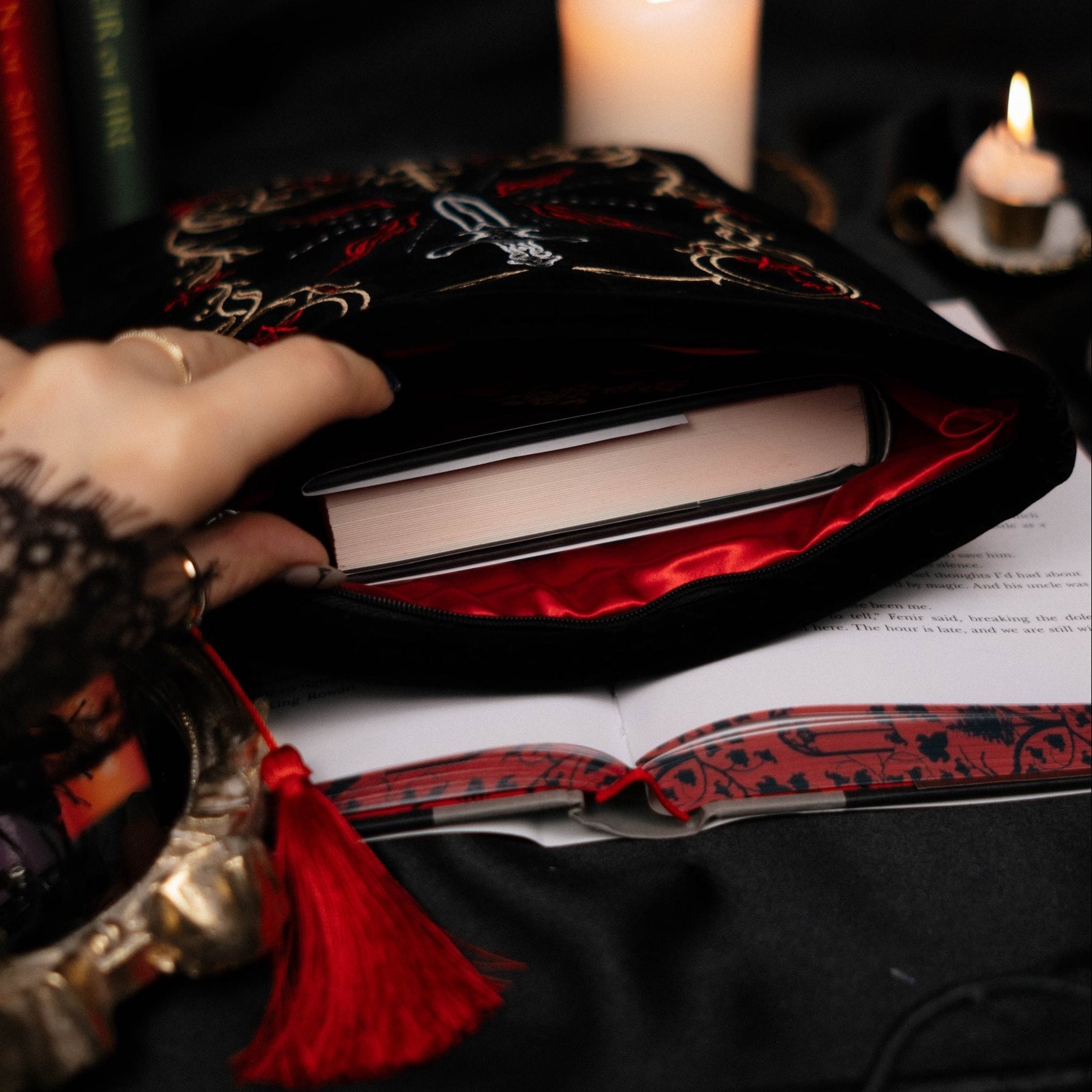 Close-up of a hand holding a red and black book sleeve open with candles and other items on a dark background