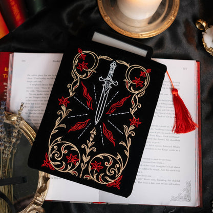Decorative kindle sleeve with a sword and tassel design on a dark surface with candles and books.