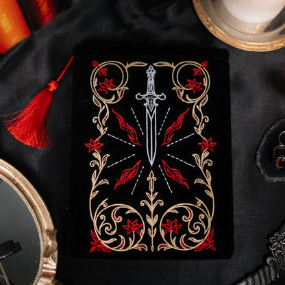 Decorative kindle pouch with a sword design on a black surface with books and a candle.
