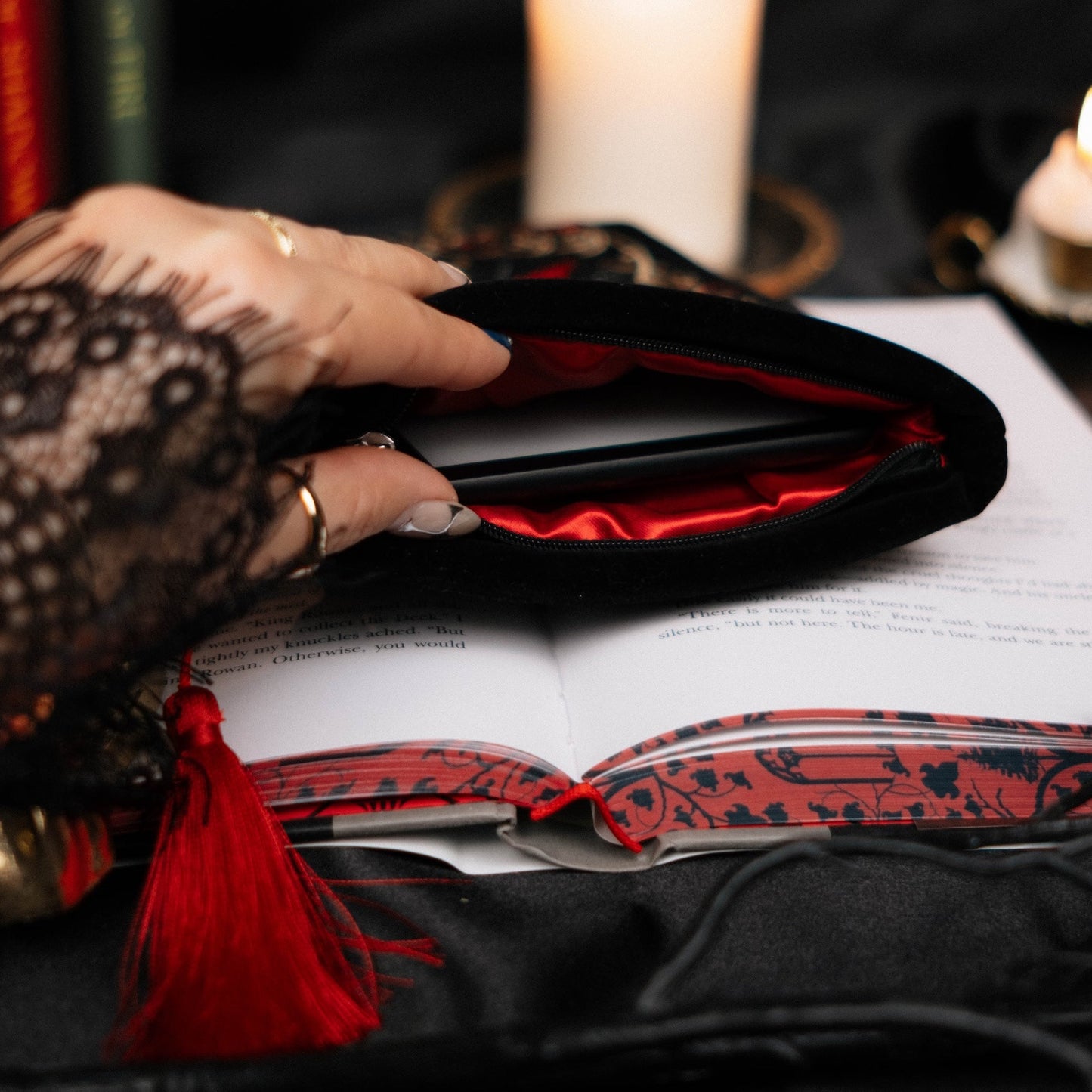 Hand reaching into a black kindle sleeve with red lining on an open book, surrounded by candles and books, against a dark background.