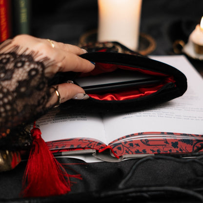 Hand reaching into a black kindle sleeve with red lining on an open book, surrounded by candles and books, against a dark background.