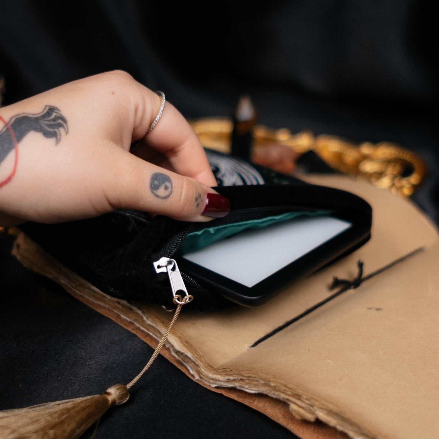 Hand with tattoo holding a small black kindle pouch over an open book with mystical items.