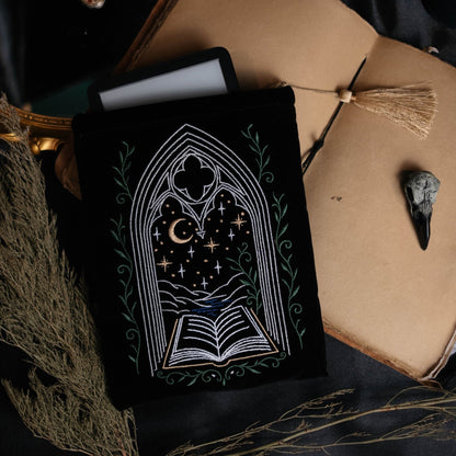 Decorative kindle pouch featuring a gothic window design surrounded by a beige book, on a dark fabric background.